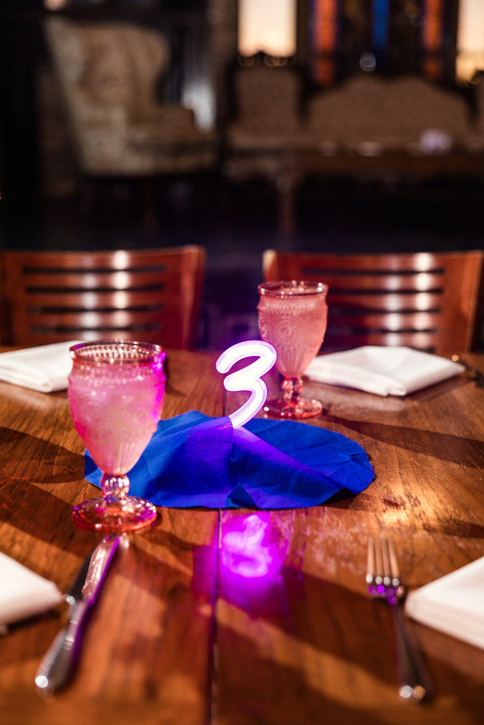 A wooden table set for four with pink drinks and a glowing number 3 centerpiece on blue fabric for Salvage One wedding reception