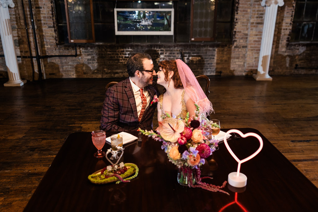 Bride and grooom in wedding attire sits at a sweetheart table, leaning in for a kiss during their Salvage One wedding reception