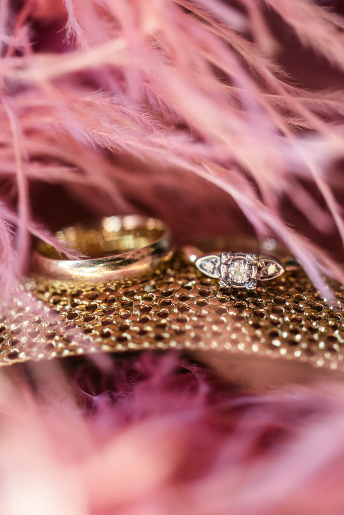Two gold rings, one with a diamond, rest on a textured surface surrounded by soft pink feathers for Salvage One wedding celebration