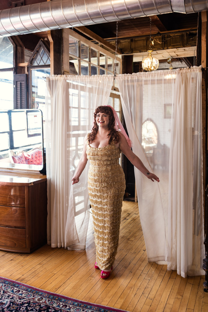 Bride in a gold, textured dress and pink heels smiles while stepping through white curtains during her first look at Salvage One