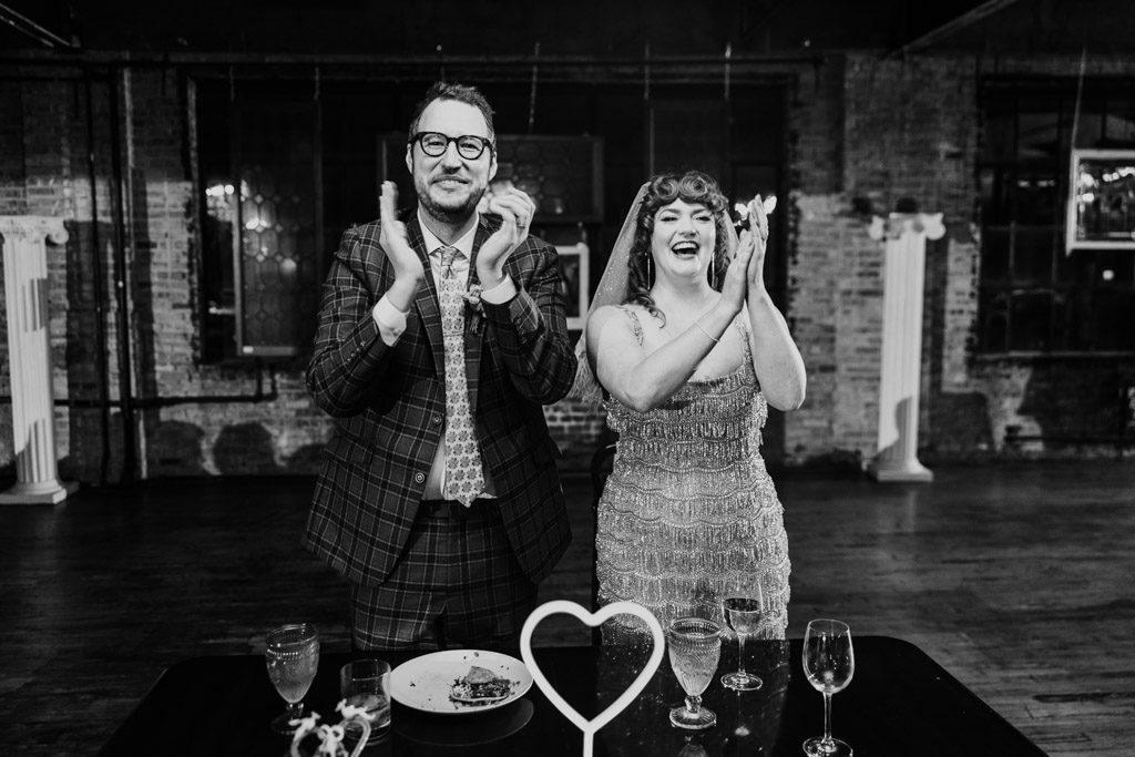 Black and white photo of bride and groom standing and clapping behind a table with drinks and a heart decoration during their Salvage One wedding reception