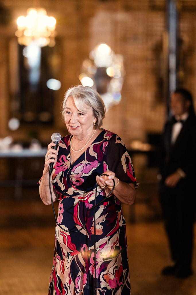 Guest in a floral dress speaks into a microphone at Salvage One wedding reception, illuminated by warm lighting