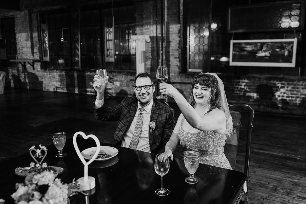 Black and white photo of newlyweds at a table raising glasses in a toast during their Salvage One wedding reception