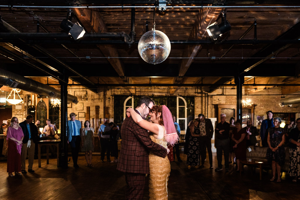 Newlywed couple shares their first dance under a disco ball at their Salvage One wedding reception, surrounded by guests