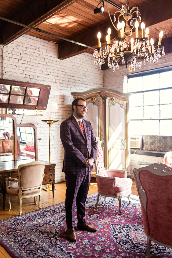 Groom in a plaid suit smiles at the bride while standing in a vintage, sunlit room with elegant furniture during his first look at Salvage One