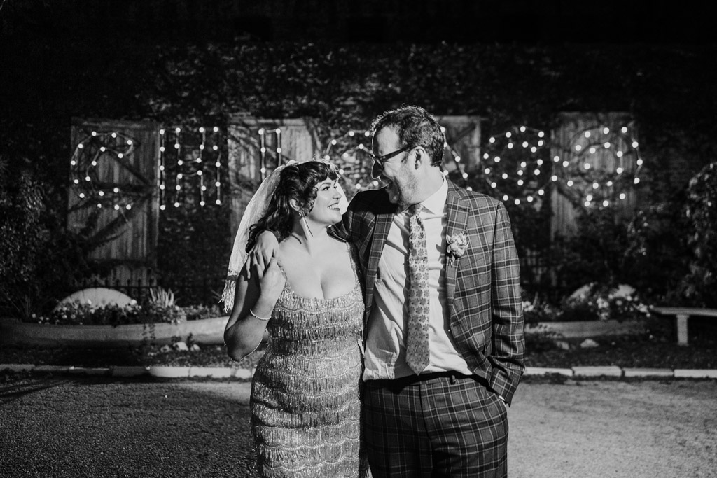 Black and white photo of bride and groom smiling at each other outside Salvage One at night, with a lit 