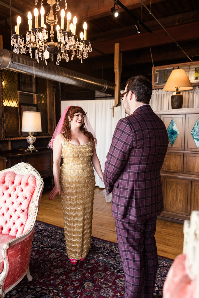 A bride in a gold dress and pink veil smiles at a groom in a plaid suit during their first look inside Salvage One's bridal suite