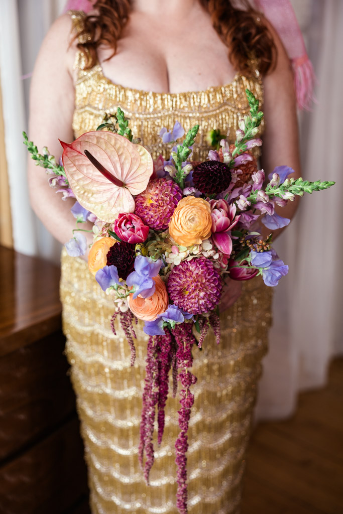 Detail photo of bride in a gold dress holding her colorful bouquet with pink, orange, and purple flowers before her Salvage One wedding