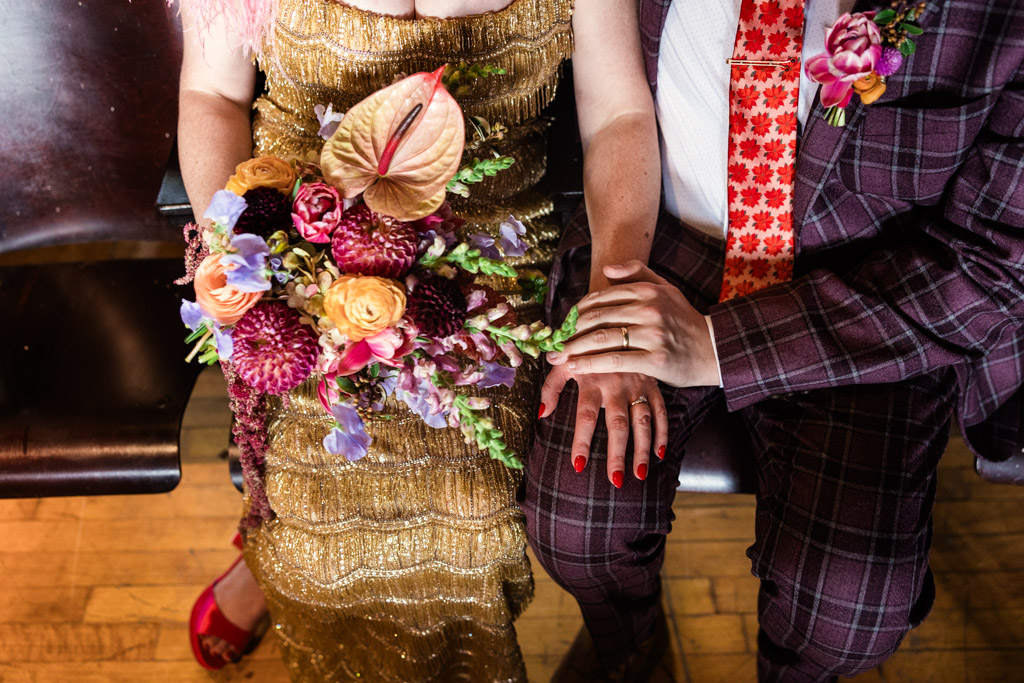 Bride and groom in colorful outfits hold hands, showing rings and a vibrant bouquet, sitting closely together before their Salvage One wedding