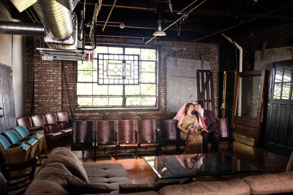 Bride and groom in colorful attire kiss while sitting on old theater seats inside Salvage One wedding venue