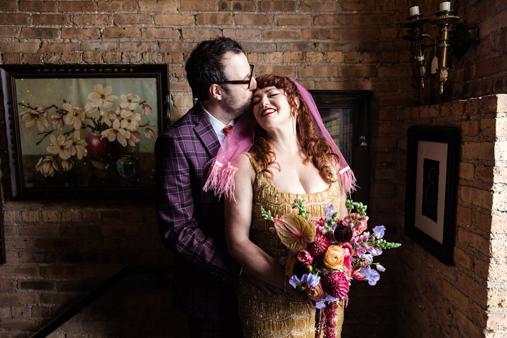 Groom in a plaid suit kisses bride in a gold dress on the forehead while she holds a colorful bouquet before their Salvage One wedding