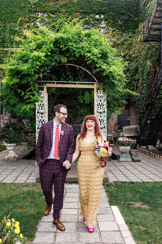 Bride and groom in colorful attire walk hand-in-hand in a garden under a leafy archway outside Salvage One