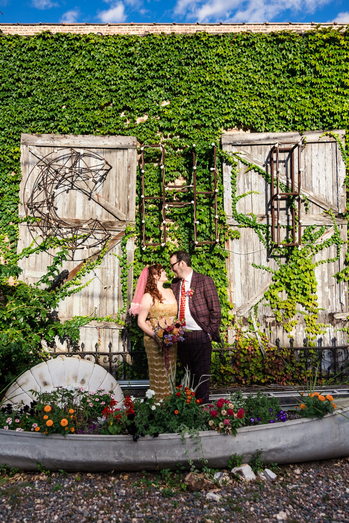 Bride and groom in colorful outfits stand in front of an ivy-covered wall and colorful garden, gazing at each other before their Salvage One wedding