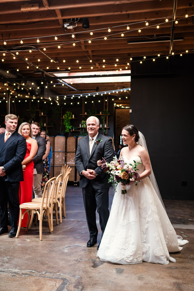 Bride in a white dress walks down the aisle with her father under string lights in The Arbory Chicago’s wedding ceremony space