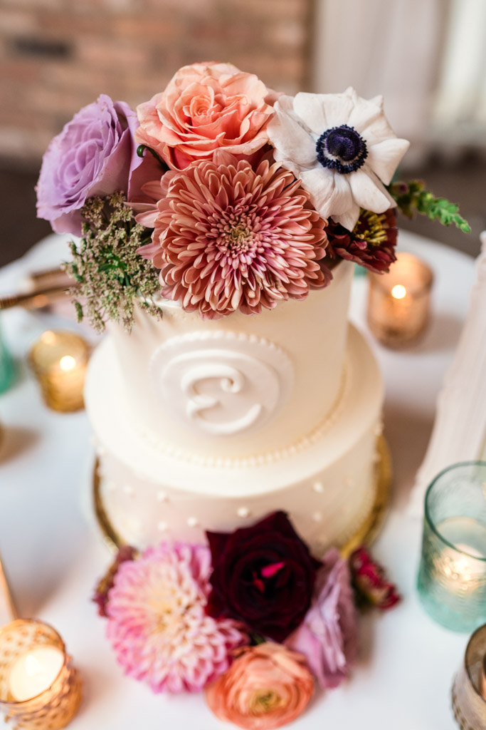 White tiered cake decorated with large pink and purple flowers, surrounded by candles and glass holders for wedding reception at The Arbory Chicago
