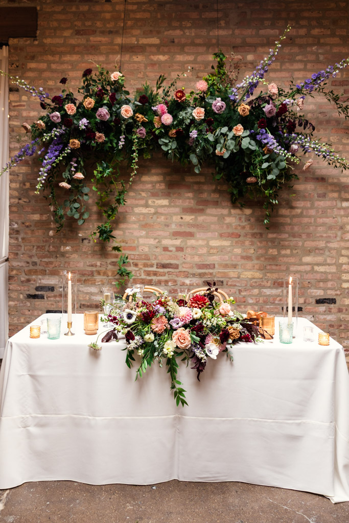 Sweetheart table with floral centerpieces and candles in front of a brick wall, beneath a large hanging flower arrangement for wedding reception at The Arbory Chicago