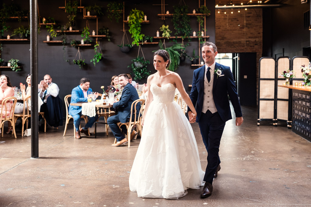 Bride and groom holding hands enter The Arbory Chicago wedding reception as guests seated at tables watch and smile
