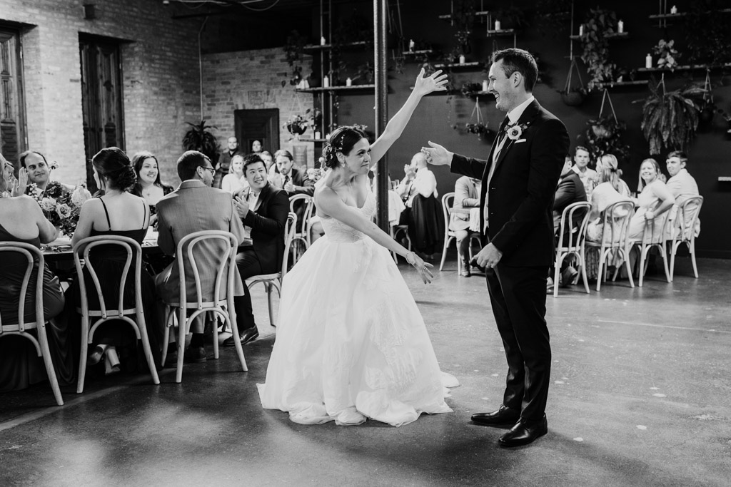 Black and white photo of bride playfully indicating the groom as they enter their wedding reception at The Arbory Chicago while guests smile and watch from tables