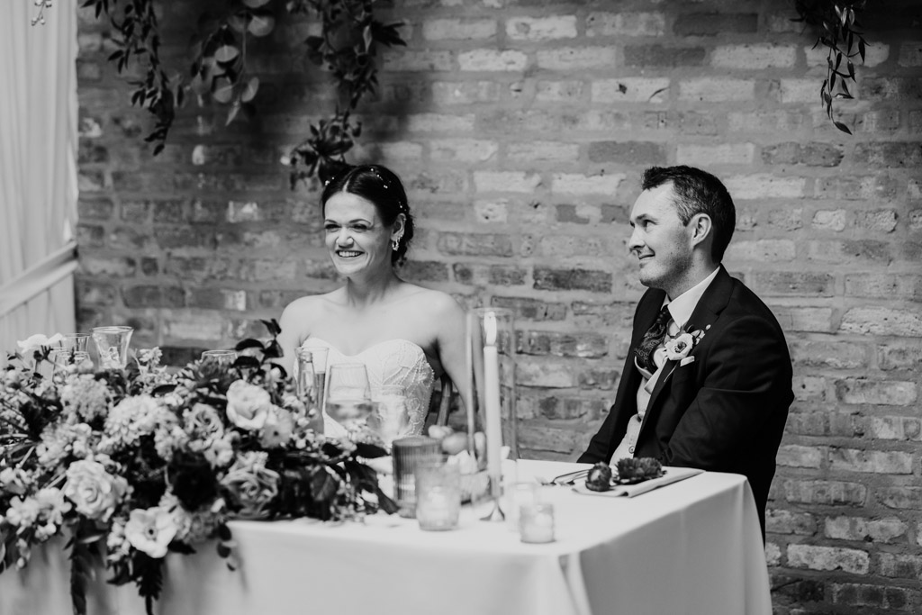 Black and white photo of bride and groom sitting smiling at the head table during their wedding reception at The Arbory Chicago