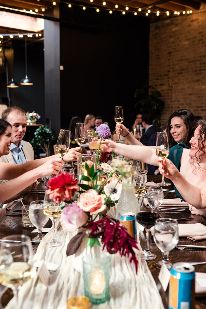 Guests raise glasses for a toast during wedding reception at The Arbory Chicago, gathered around a table decorated with flowers