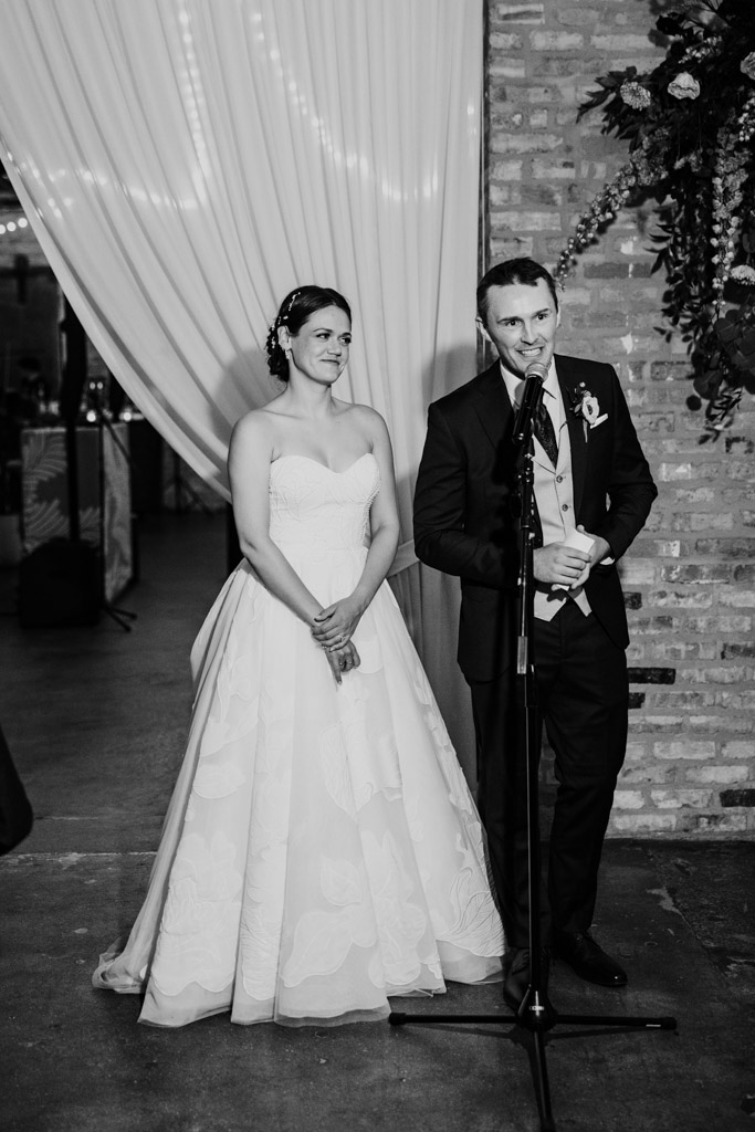 Black and white photo of bride and groom standing together at a microphone, smiling during their Arbory Chicago wedding reception