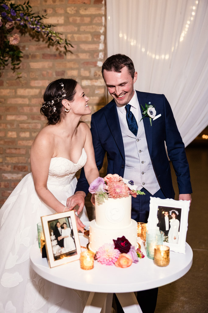Bride and groom smiling as they cut their wedding cake at The Arbory Chicago, with framed photos and flowers decorating the table