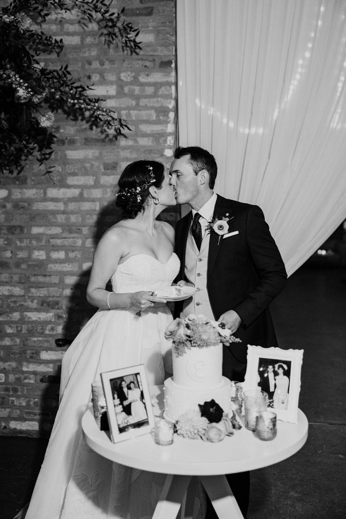 Black and white photo of bride and groom kissing after cutting their wedding cake on a small table with framed photos and candles at The Arbory Chicago