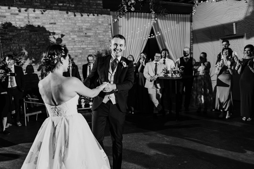 Black and white photo of bride and groom sharing their first dance, smiling, surrounded by guests at their wedding reception at The Arbory Chicago