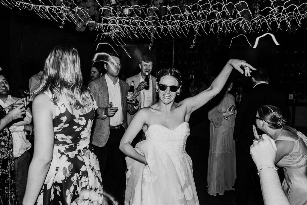 Creative black and white photo of bride in sunglasses dancing at her wedding reception at The Arbory Chicago, surrounded by guests