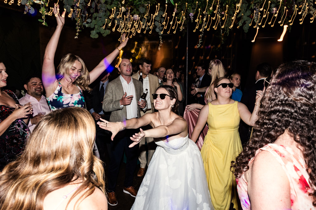 Bride in sunglasses dances with guests under string lights during her wedding reception at The Arbory Chicago
