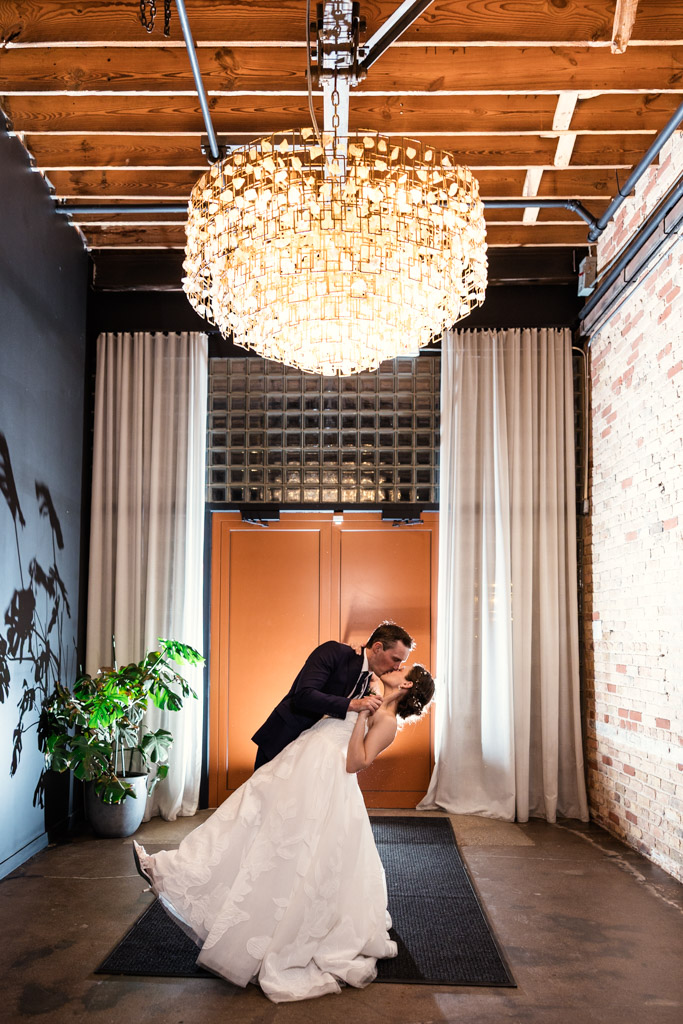 Groom dips and kisses his bride under a large chandelier during their wedding reception at The Arbory Chicago