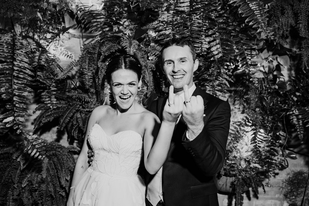 Black and white photo of happy bride and groom smiling and showing their wedding rings in front of leafy greenery at Garfield Park Conservatory