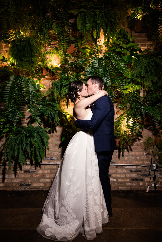 Bride and groom kiss in front of a lush green backdrop with hanging ferns at The Arbory Chicago