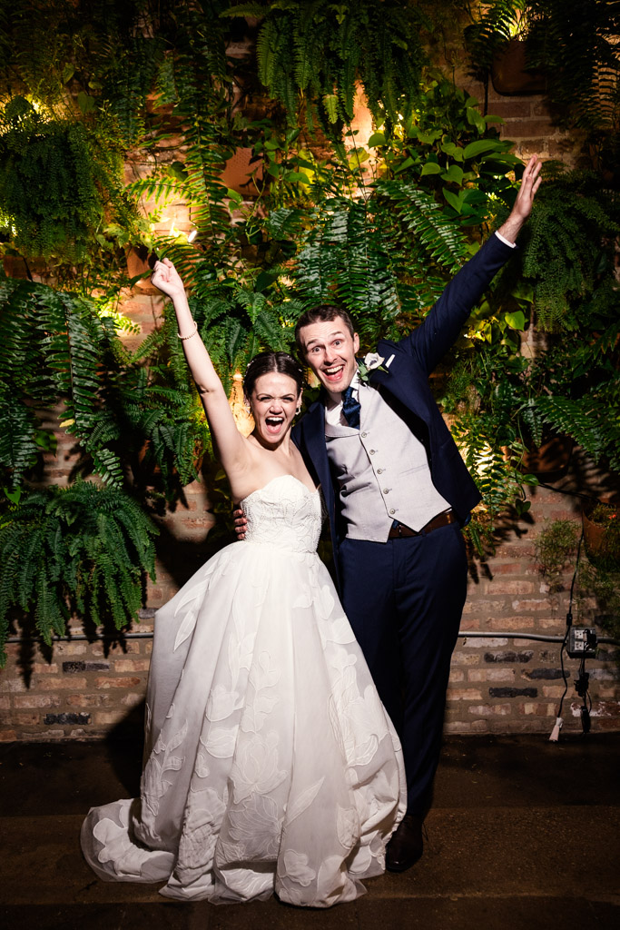 A joyful bride and groom celebrate with arms raised in front of a lush, green fern backdrop at The Arbory Chicago