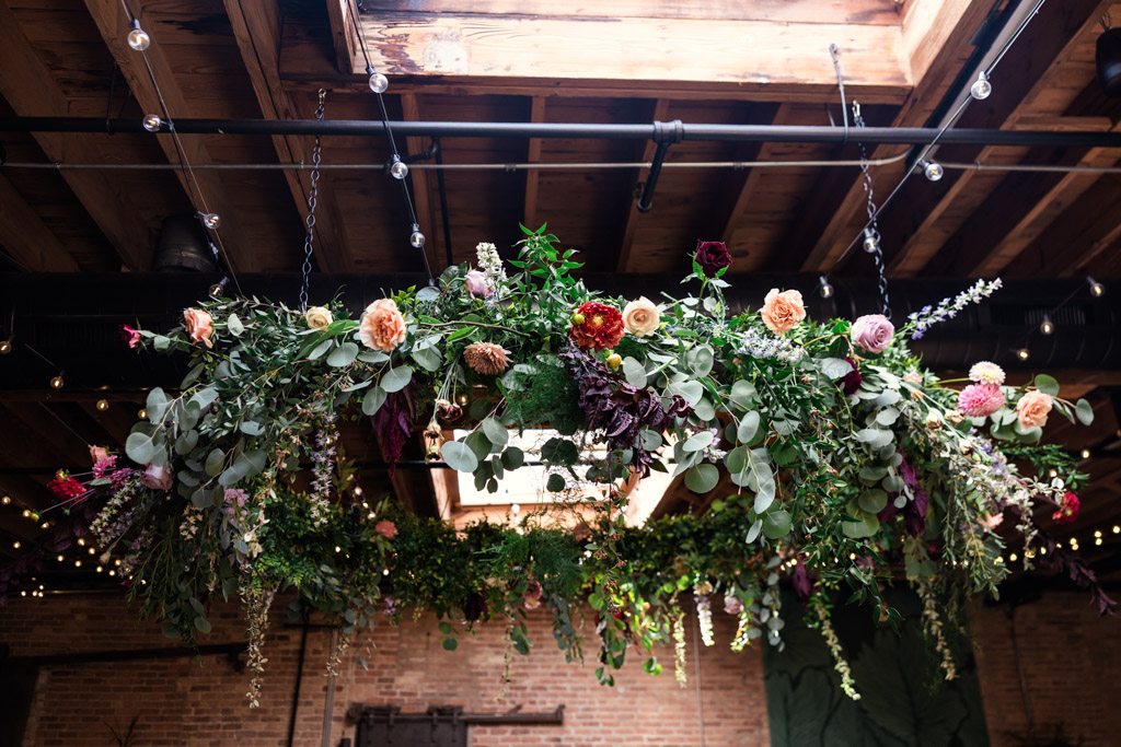 Hanging floral arrangement with greenery and colorful flowers under a wooden ceiling with string lights for wedding celebration at The Arbory Chicago