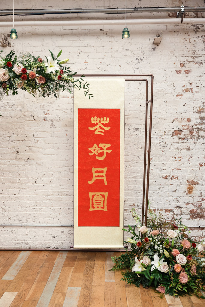 Vertical red banner with gold Chinese characters, surrounded by floral arrangements for wedding ceremony at The Joinery in Chicago