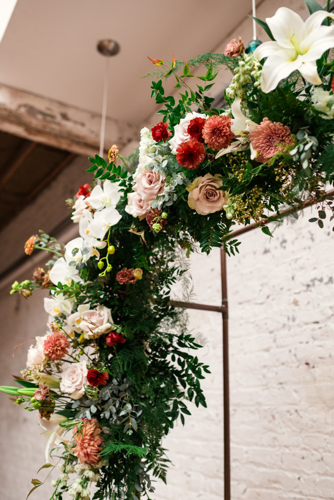 Detail photo of floral decor on arch with white, pink, and red flowers displayed against a white brick wall for wedding ceremony