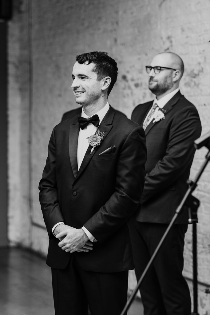 Black and white photo of groom in a tuxedo watching the bride walk down the aisle during their Joinery wedding ceremony