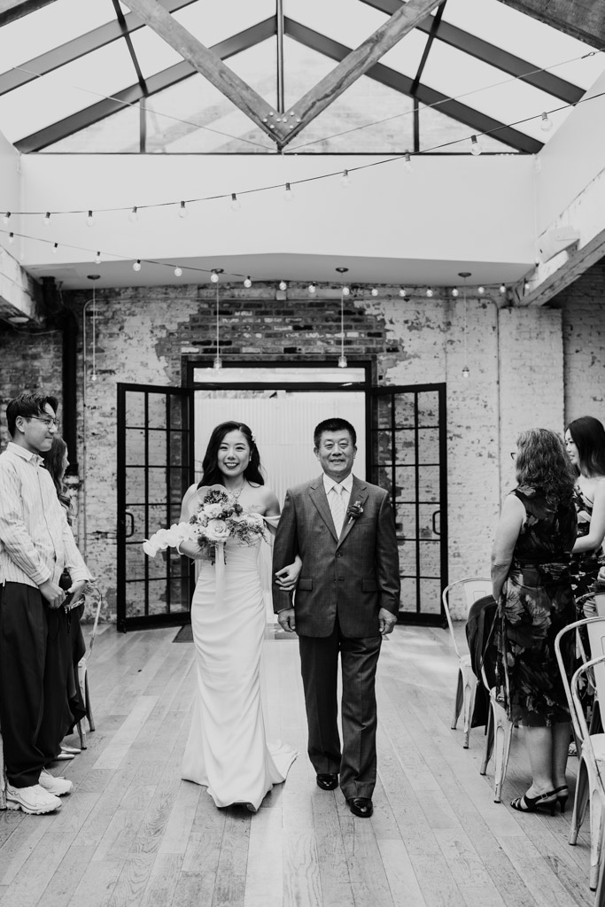 Black and white photo of bride walking down the aisle with her father in the wedding ceremony space of The Joinery in Chicago