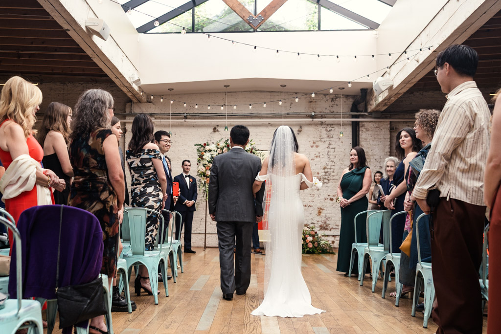 Bride and her father walk down the aisle together as guests stand and smile inside The Joinery's wedding ceremony space