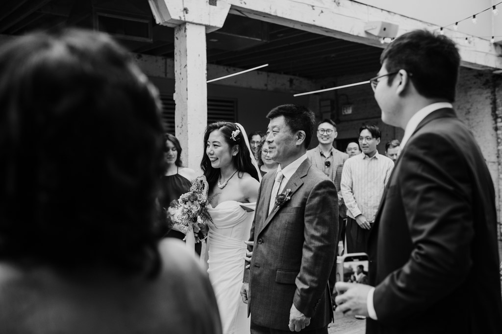 Black and white photo of bride and her father walking down the aisle during wedding ceremony at The Joinery in Chicago