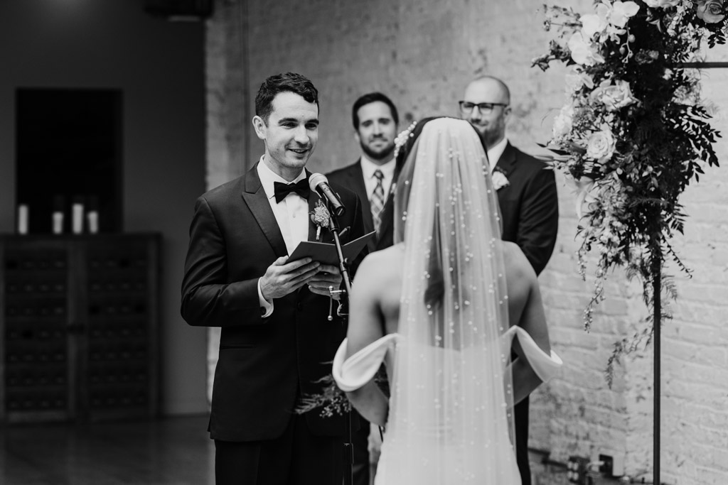 Groom reads vows to his bride at their wedding ceremony, with groomsmen standing behind them, all framed by elegant joinery details in the venue.
