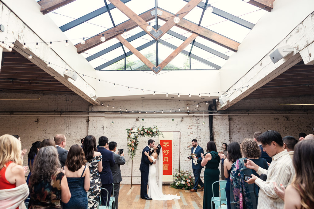 First kiss at The Joinery Chicago wedding under skylight with guests applauding in white loft venue