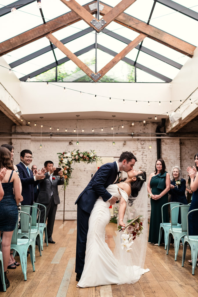 Groom dips and kisses his bride at their indoor wedding ceremony at The Joinery as guests applaud