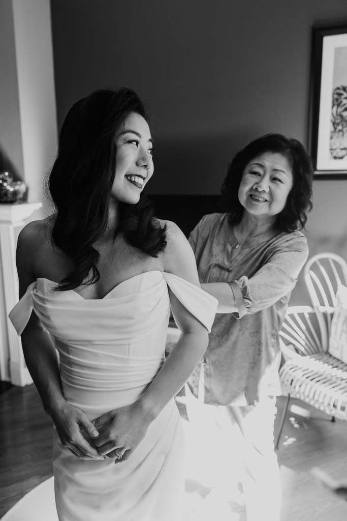 Black and white photo of bride in a white gown smiling as her mother helps her get ready before her wedding at The Joinery in Chicago