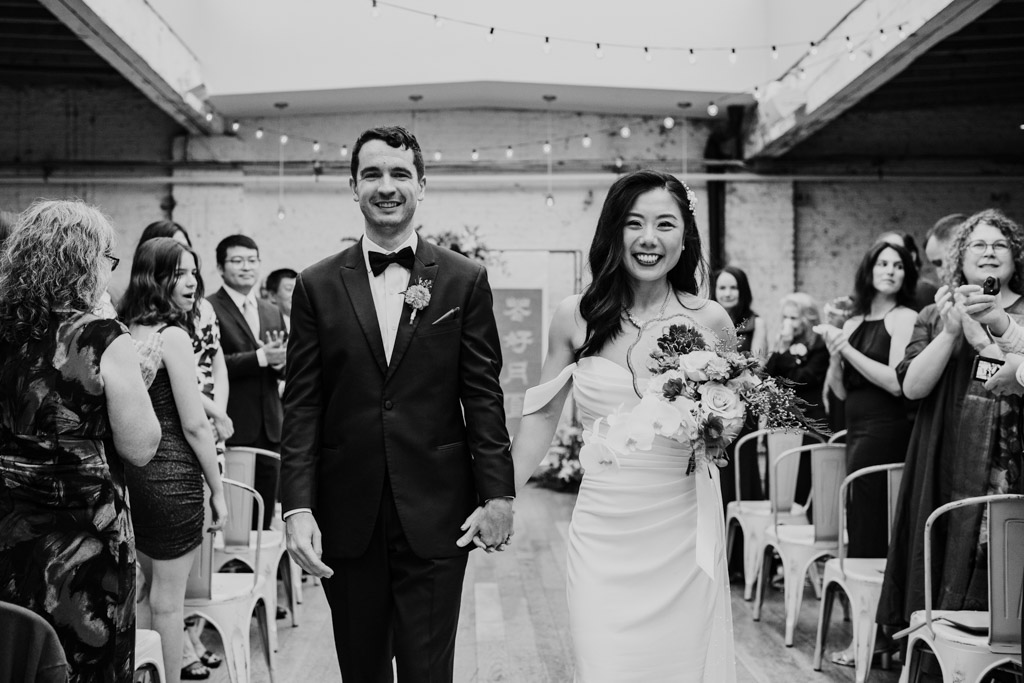 Black and white photo of bride and groom walking down the aisle smiling, surrounded by applauding wedding guests after their Joinery wedding ceremony