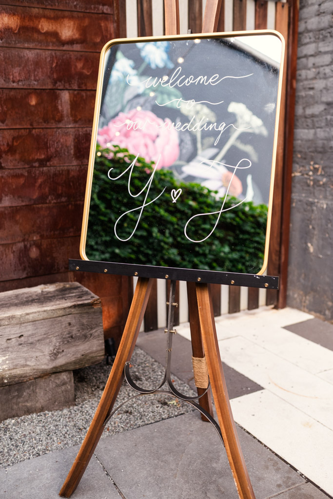 Mirrored welcome sign on an easel, flowers reflected in the glass, for wedding reception at The Joinery in Chicago