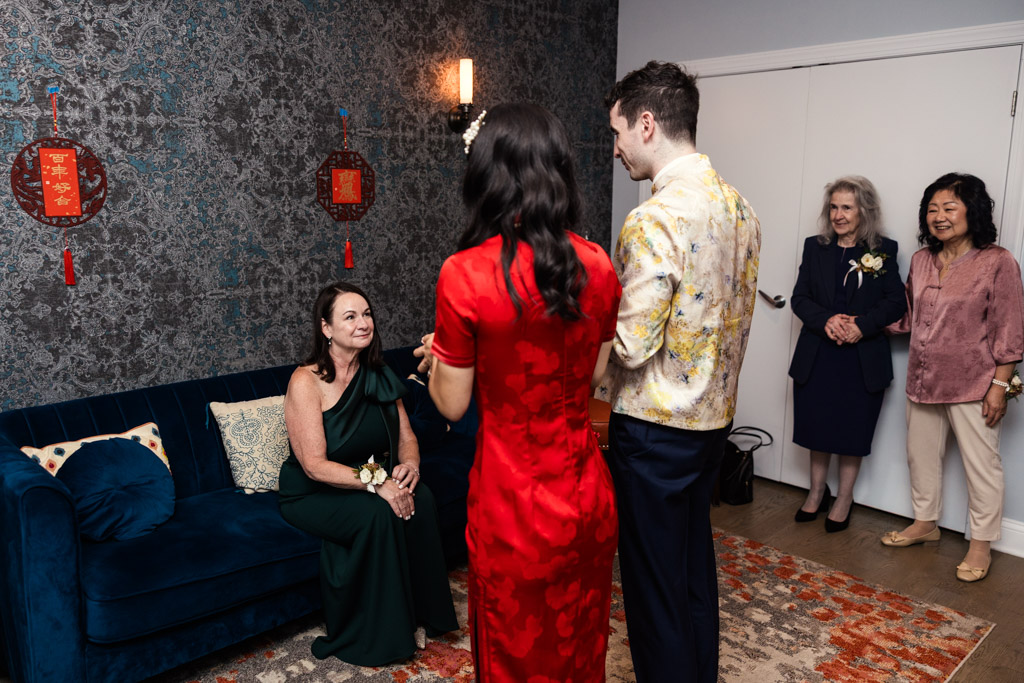 A couple in traditional attire greets a woman, while others watch in a decorated room