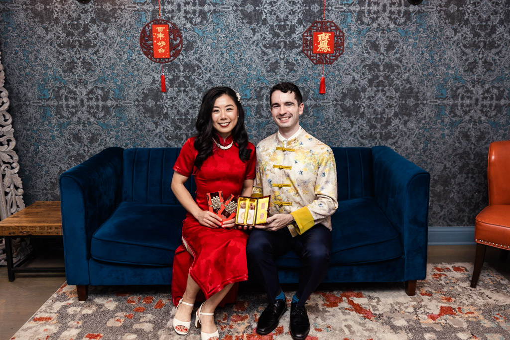 Newlywed couple in traditional Chinese attire sits on a blue couch, smiling and holding festive items for Lunar New Year