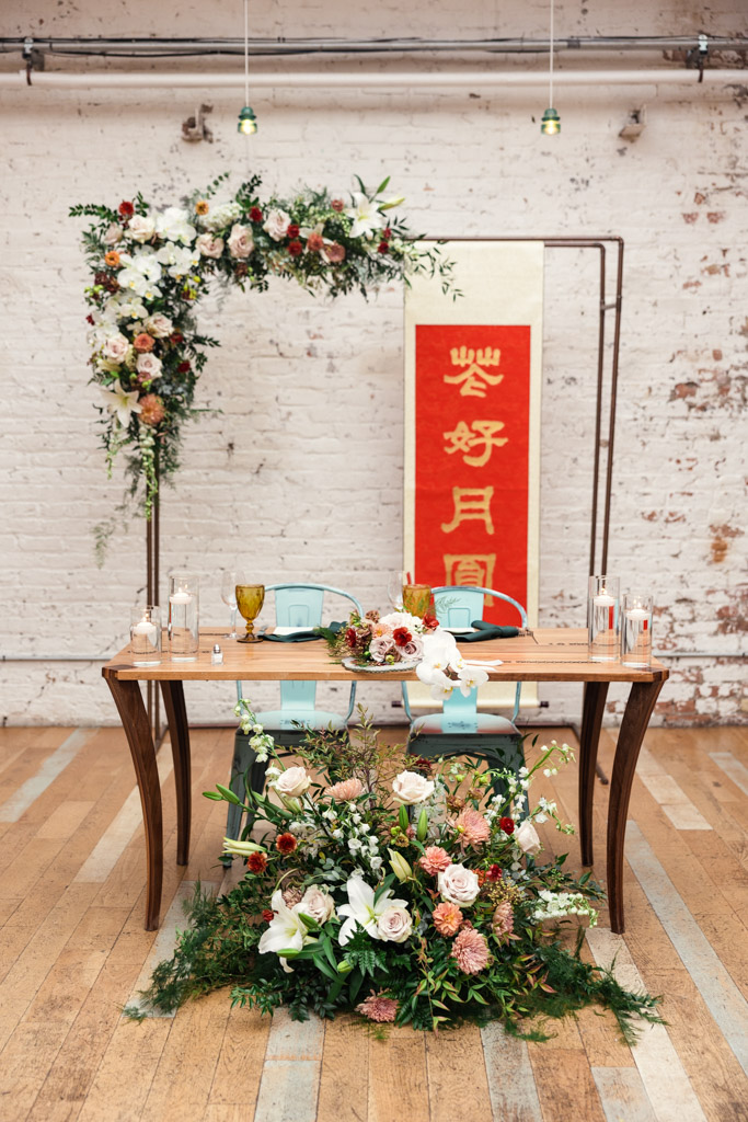 A wedding sweetheart table with flowers and a red Chinese banner is set against a white brick wall, for wedding reception at The Joinery in Chicago